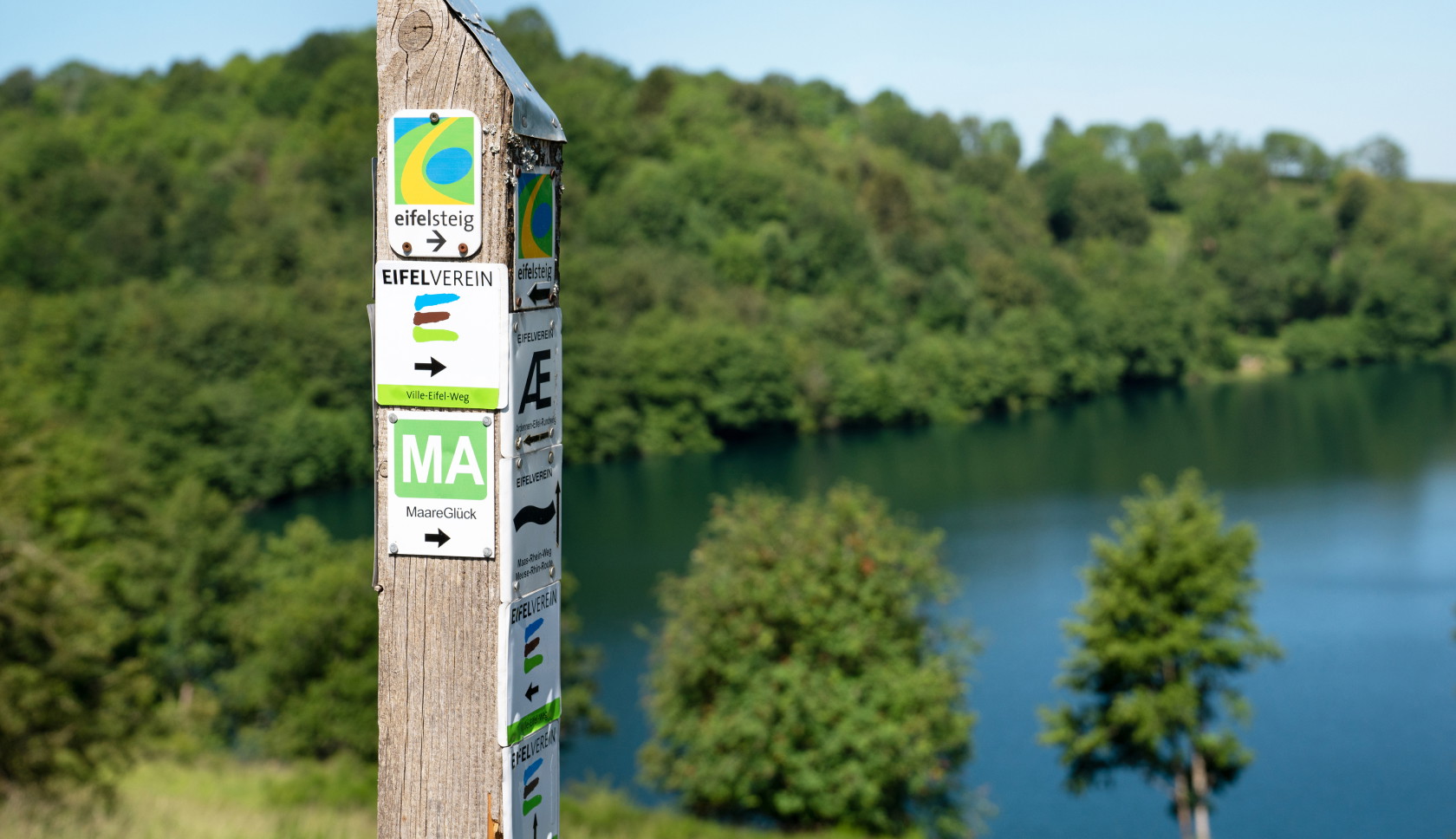 Routepaal van de Eifelsteig met uitzicht op een meer en groene heuvels