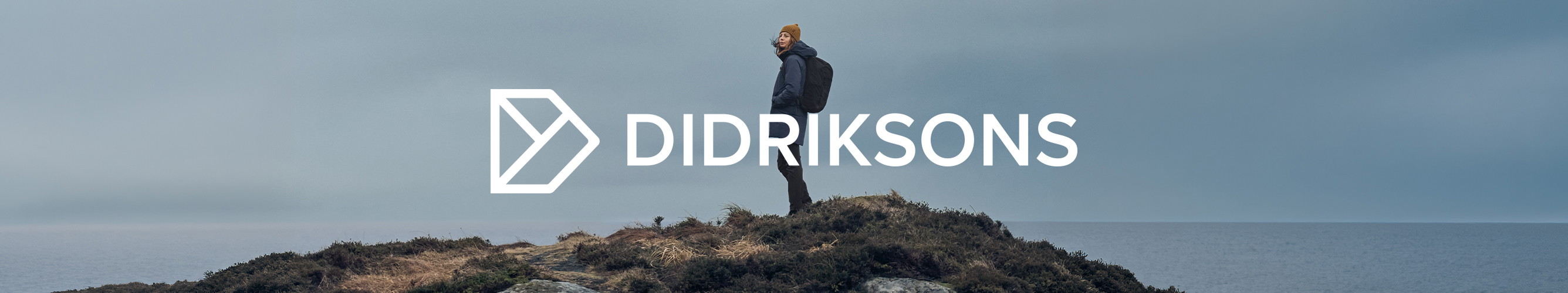 Wandelaar in Didriksons-outdoorkleding staat op een rotsachtige kust, met het Didriksons-logo centraal in beeld.