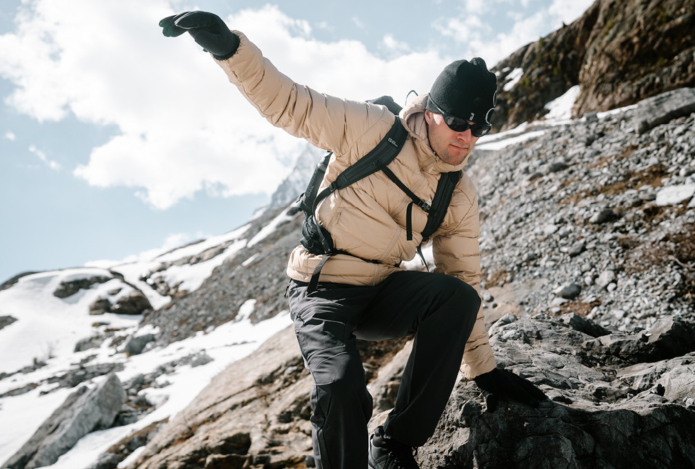 Man met beige Jack Wolfskin outdoorjas en rugzak wandelt over rotsachtig terrein in de bergen